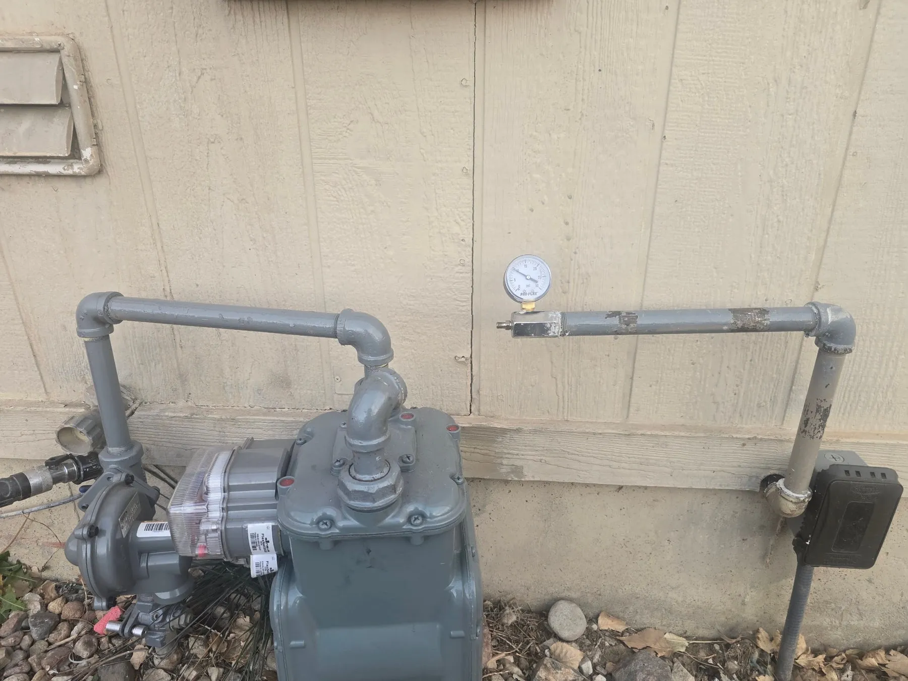 An outside gas meter and gray gas pipe against house siding, with a pressure gauge on the line