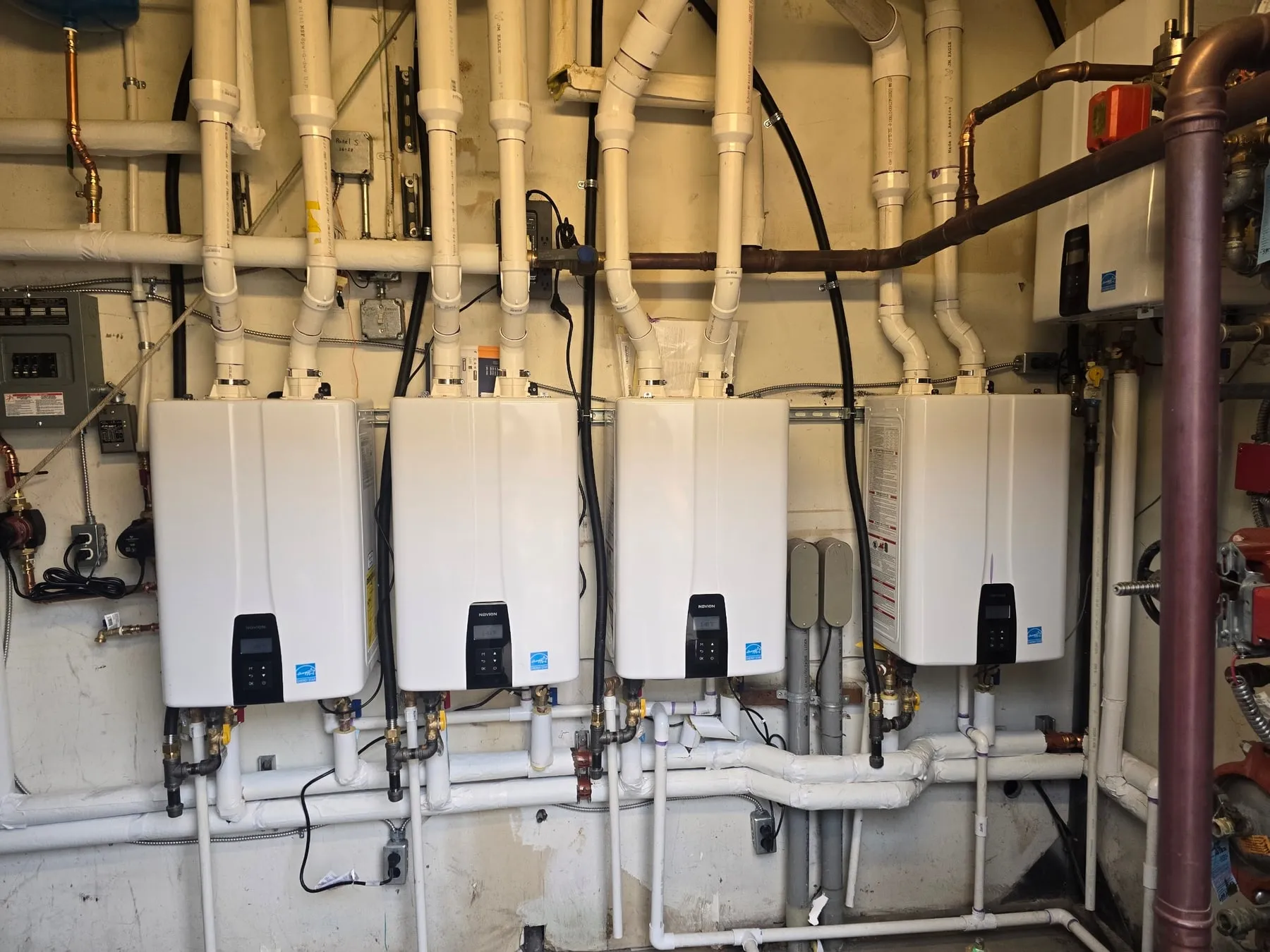 Four white tankless water heaters mounted side by side on a mechanical room wall, with their venting and water lines