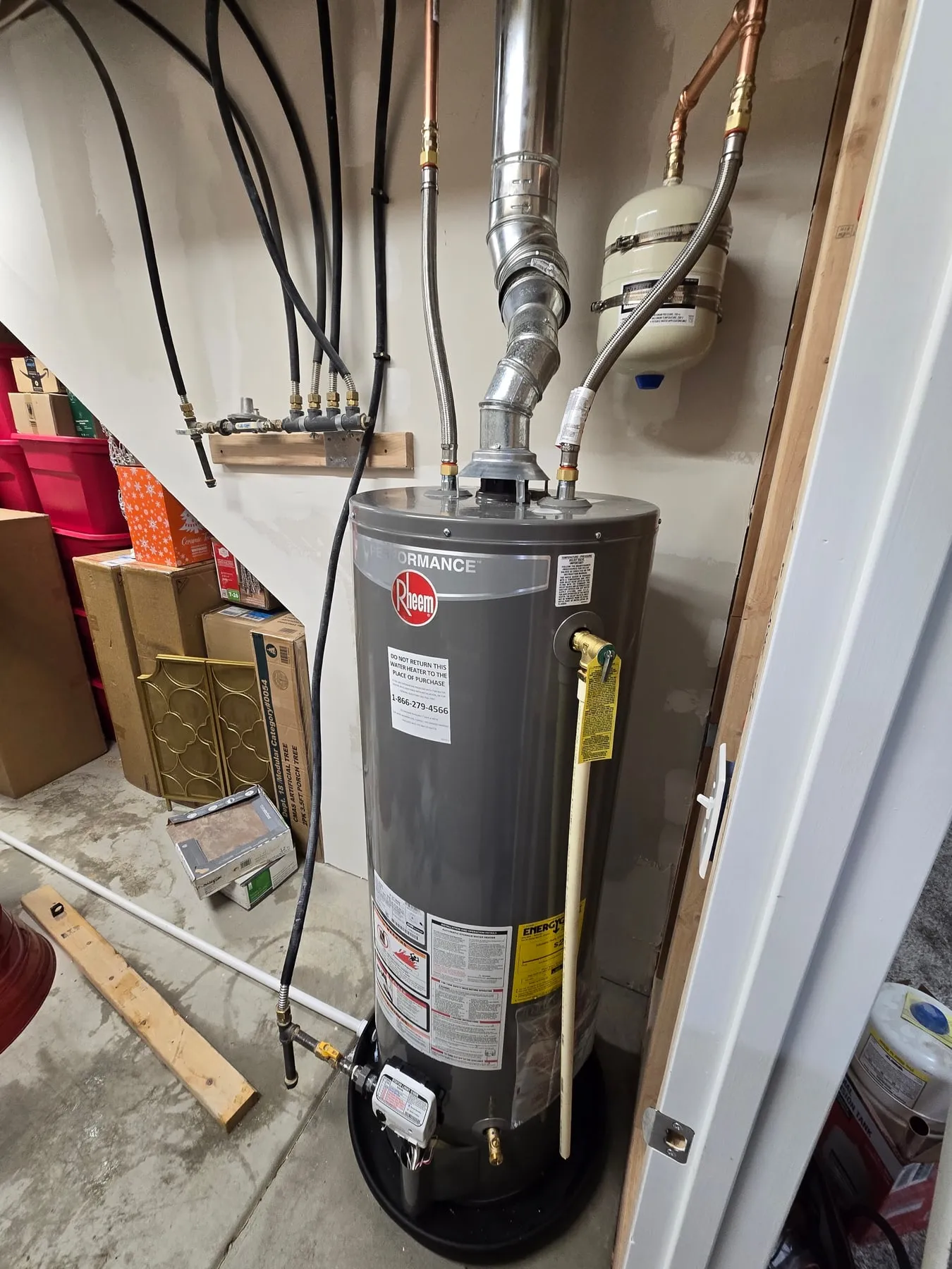 A new gray Rheem gas water heater on a drain pan in an under-stair storage room, with its flue and expansion tank